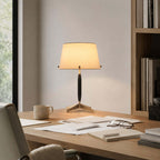 A Clérachic table lamp lit on a pale wood desk, beside a mug, loose papers and a pair of glasses, with a bookshelf behind.
