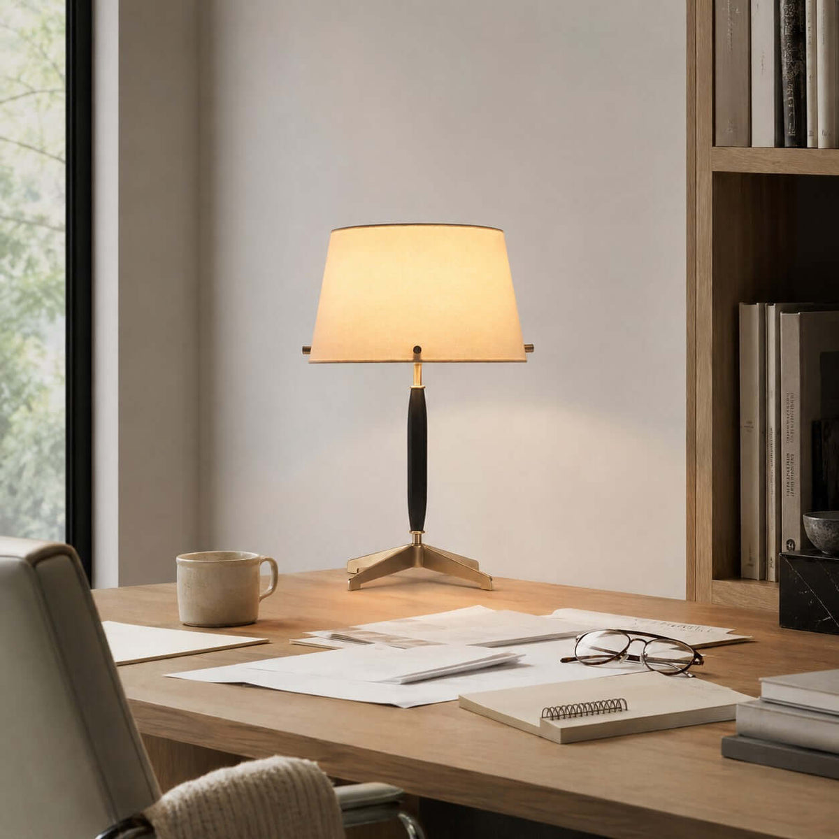 A Clérachic table lamp lit on a pale wood desk, beside a mug, loose papers and a pair of glasses, with a bookshelf behind.