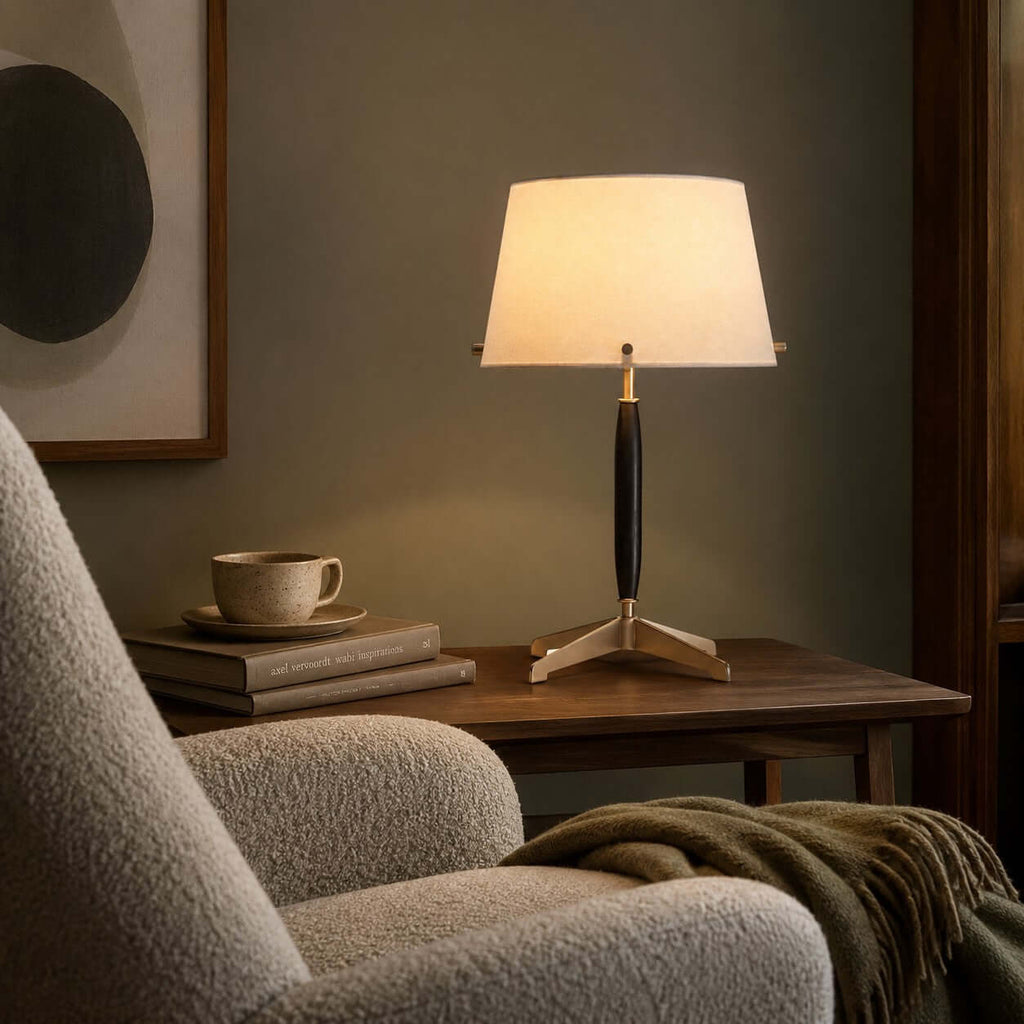Beside a boucle armchair and a green throw, this Clérachic table lamp lights a small dark wood side table with a cup and books.