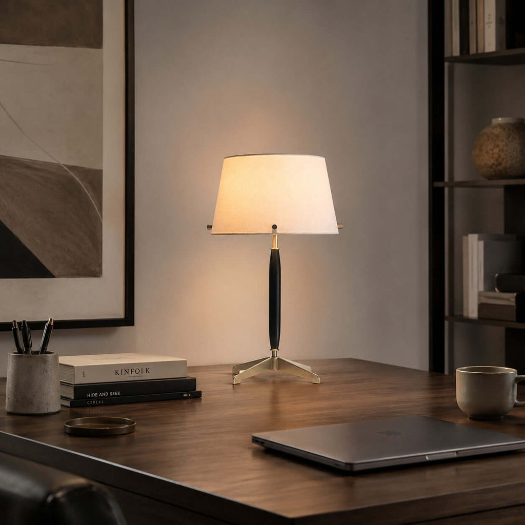 This Clérachic table lamp on a dark wood desk with a closed laptop, a pen pot and books, a framed print on the wall behind.