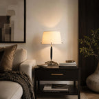 Lit Clérachic table lamp on a drawered bedside table next to a cream armchair, a folded throw and a leafy branch in a vase.