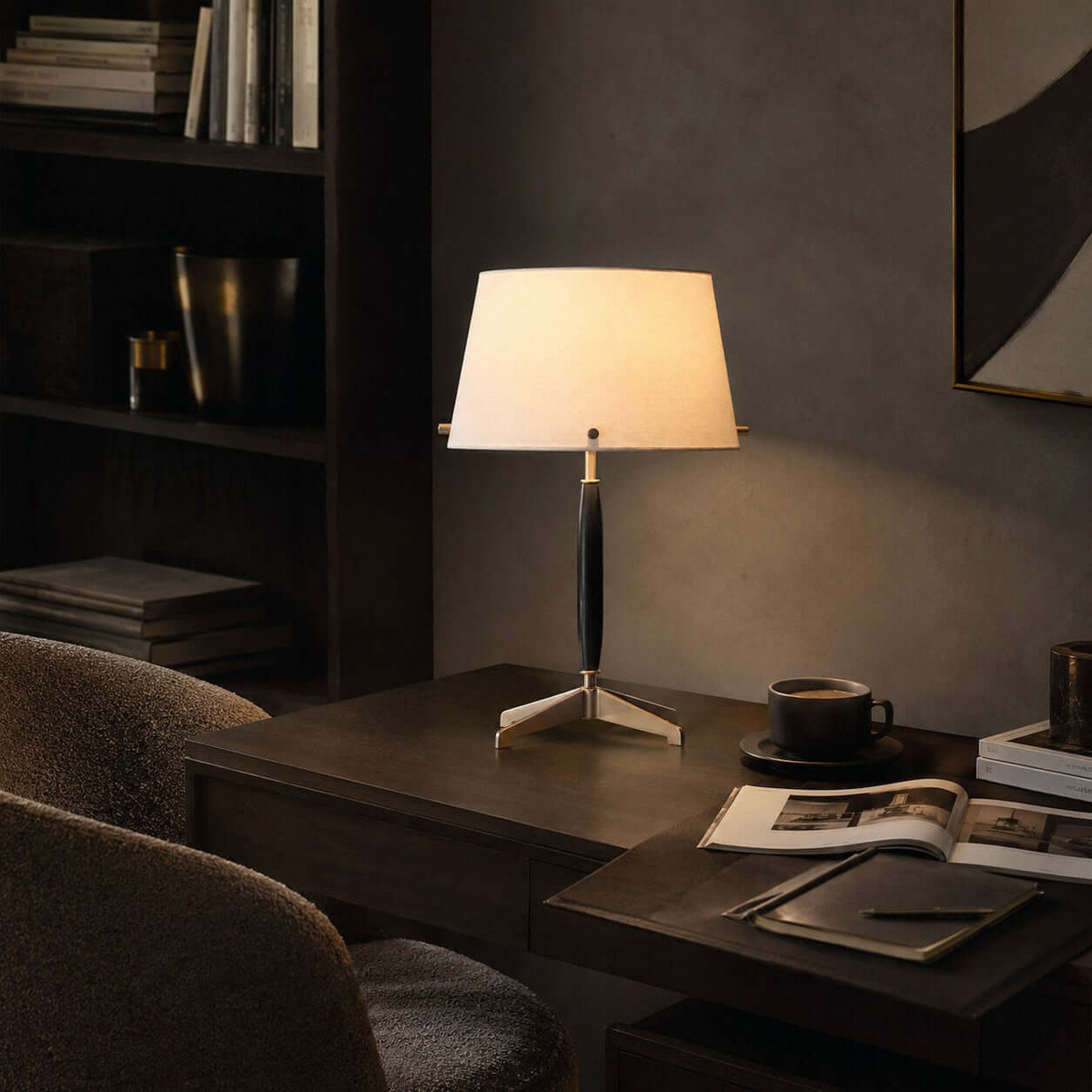 A Clérachic table lamp on a dark desk beside a coffee cup and an open magazine, with a boucle chair and shelves behind.