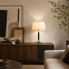 The off-white shade of this Clérachic table lamp glows on a walnut console, with a framed print, a wooden bowl and a leafy branch.