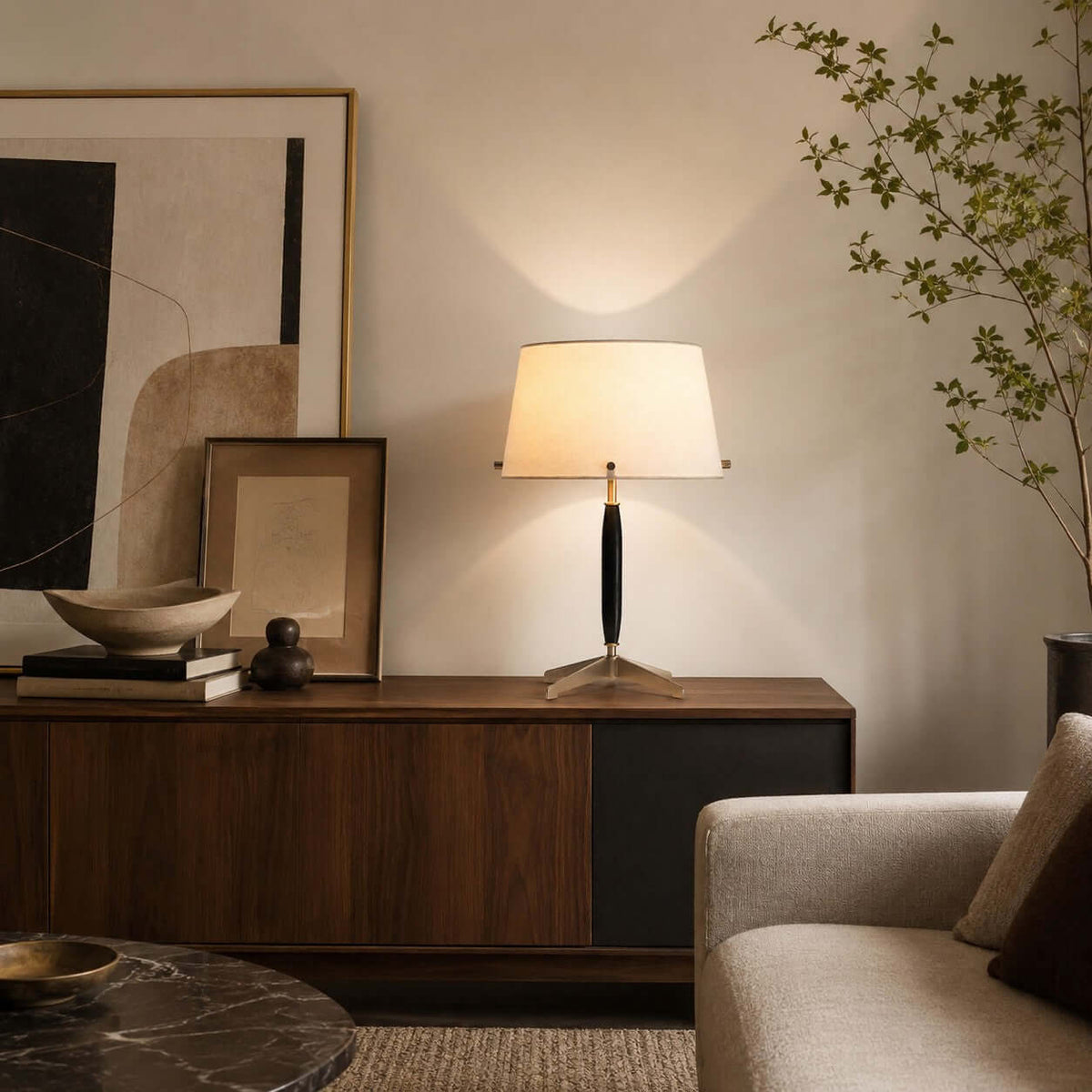 The off-white shade of this Clérachic table lamp glows on a walnut console, with a framed print, a wooden bowl and a leafy branch.