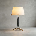 This Clérachic table lamp, unlit, stands on a polished concrete floor against a grey plaster wall, showing its dark stem and brass feet.