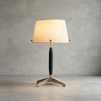 This Clérachic table lamp, unlit, stands on a polished concrete floor against a grey plaster wall, showing its dark stem and brass feet.