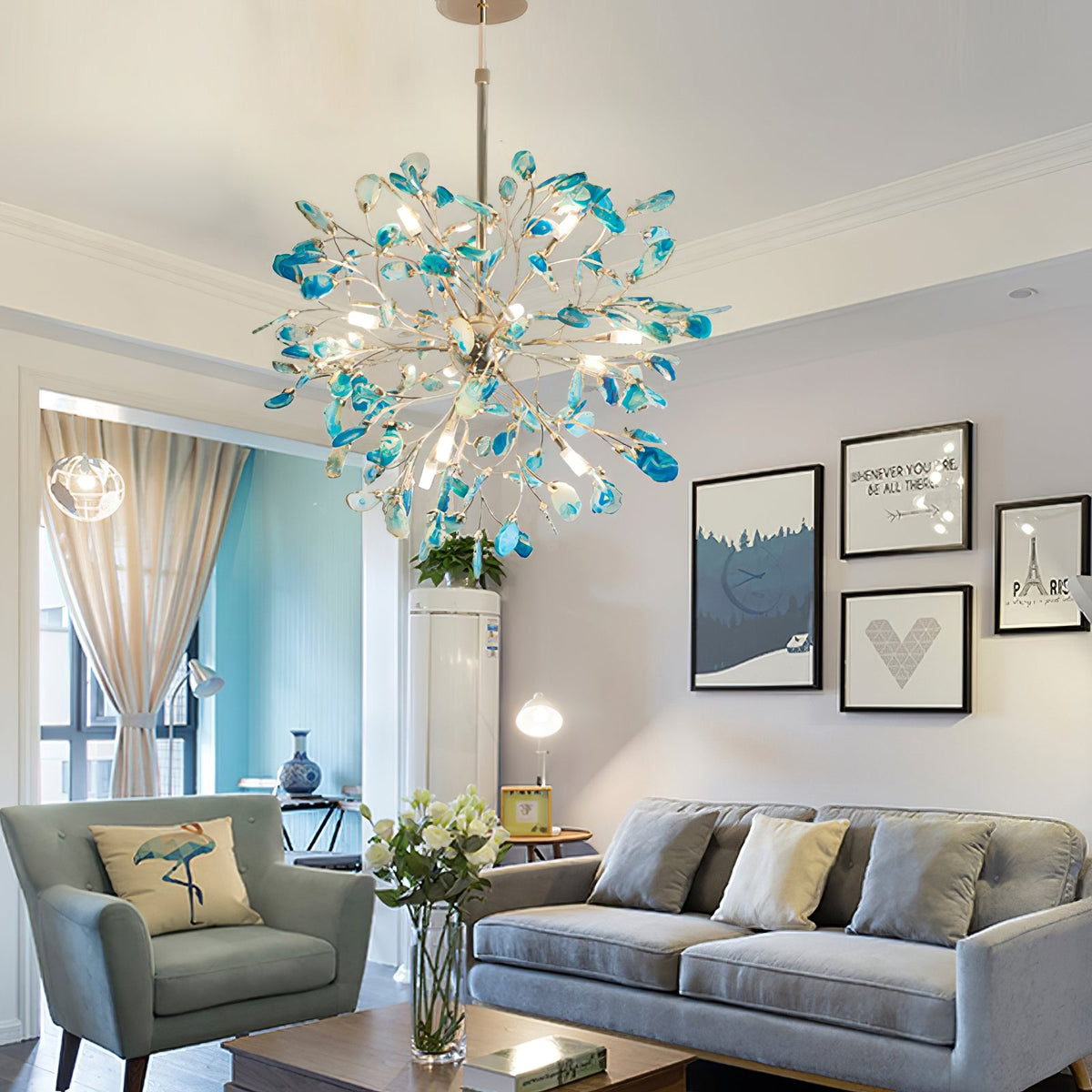 Blue-green agate Clérachic chandelier above a living room with a grey sofa, a blue armchair and framed prints on the wall.