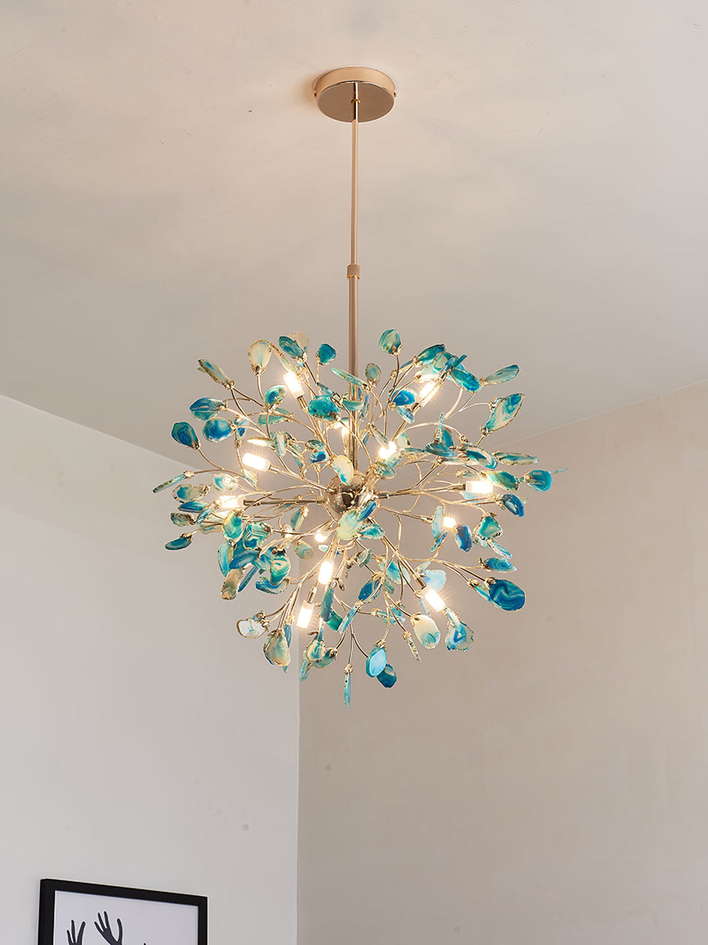 Teal agate Clérachic chandelier lit above a pale wall, with a framed picture just visible at the lower edge.