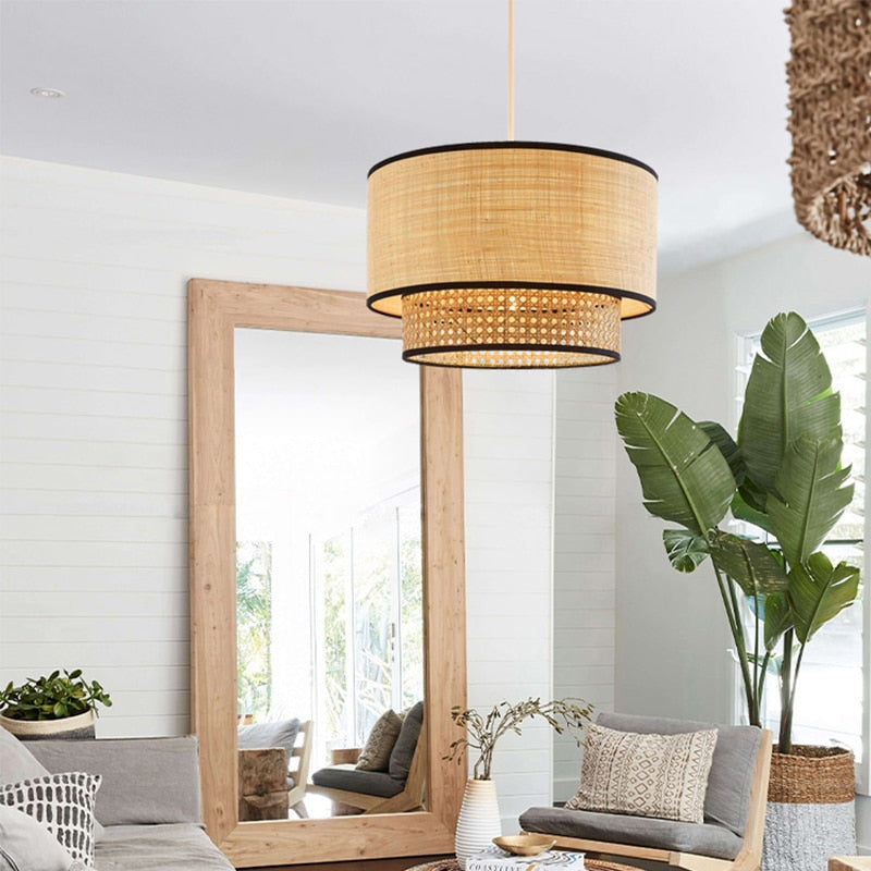 Clérachic raffia pendant lamp lit in a bright living room, beside a leaning wood-framed floor mirror and a large banana-leaf plant.
