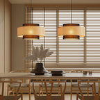 Two Clérachic pendants with woven rattan drums hang above a pale wooden table, in front of wide slatted blinds.