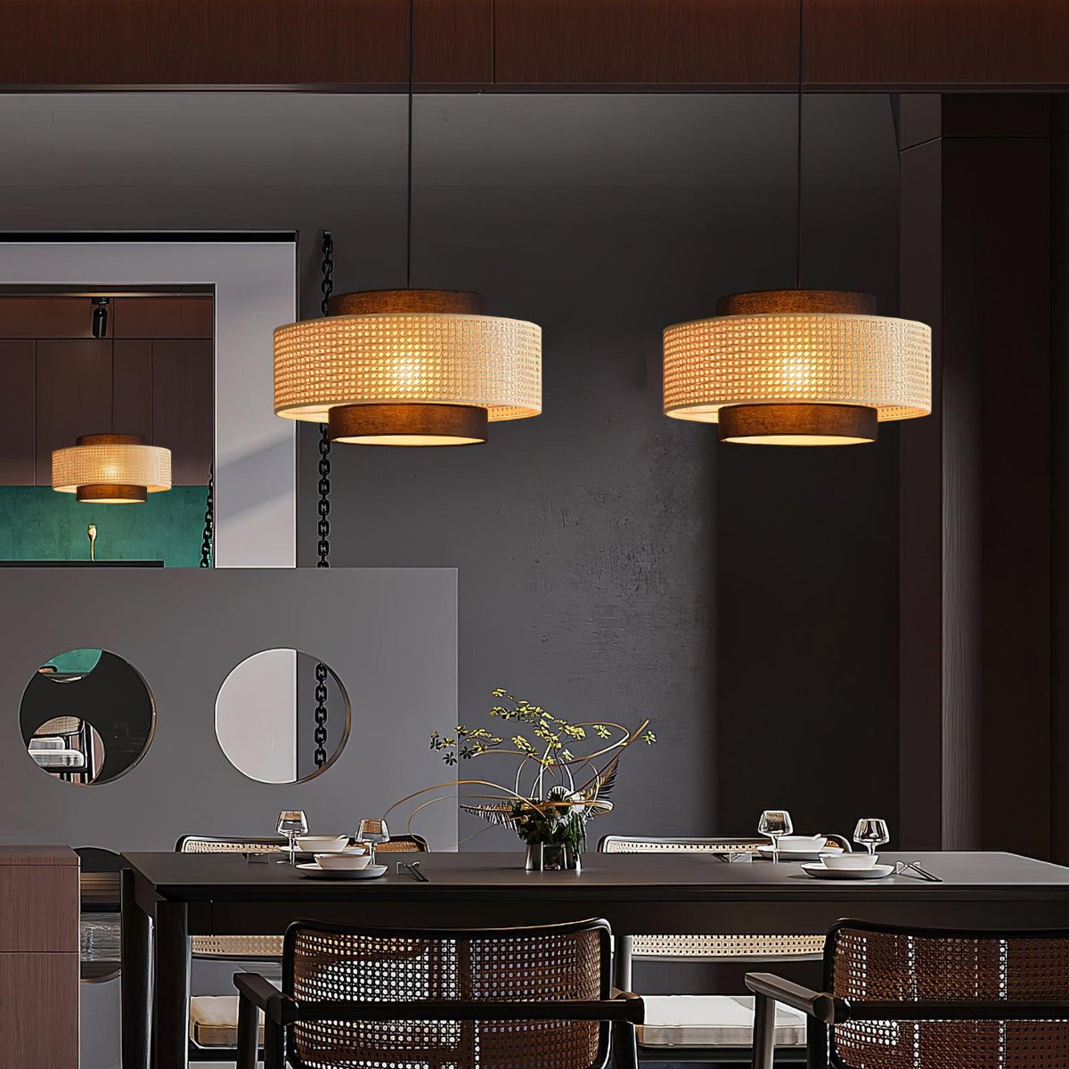 Two rattan Clérachic pendants hang above a dark dining table with woven-back chairs and a vase of branches.
