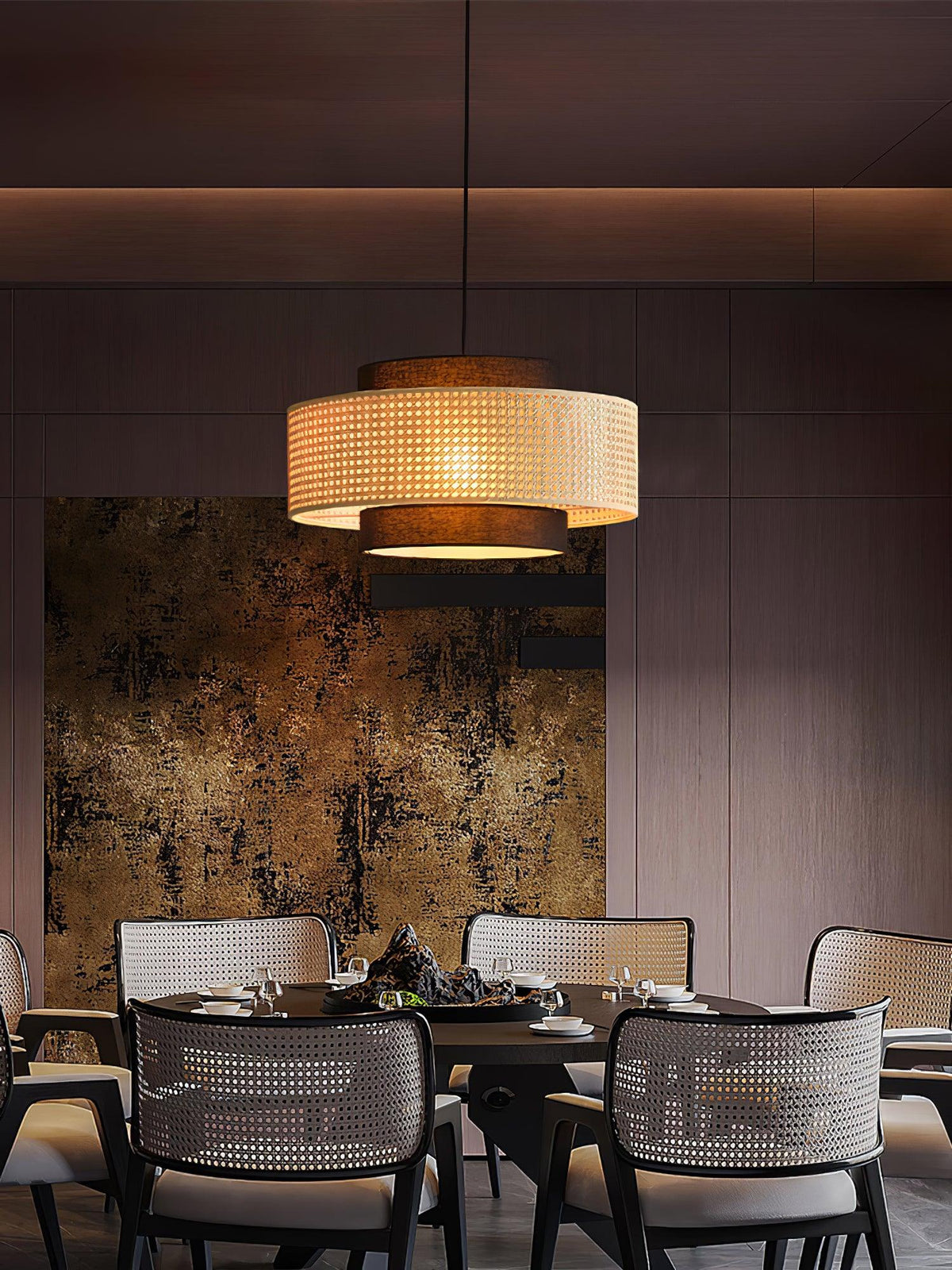A rattan Clérachic pendant glows above a table with cane-back chairs, in front of a gold-toned abstract wall panel.