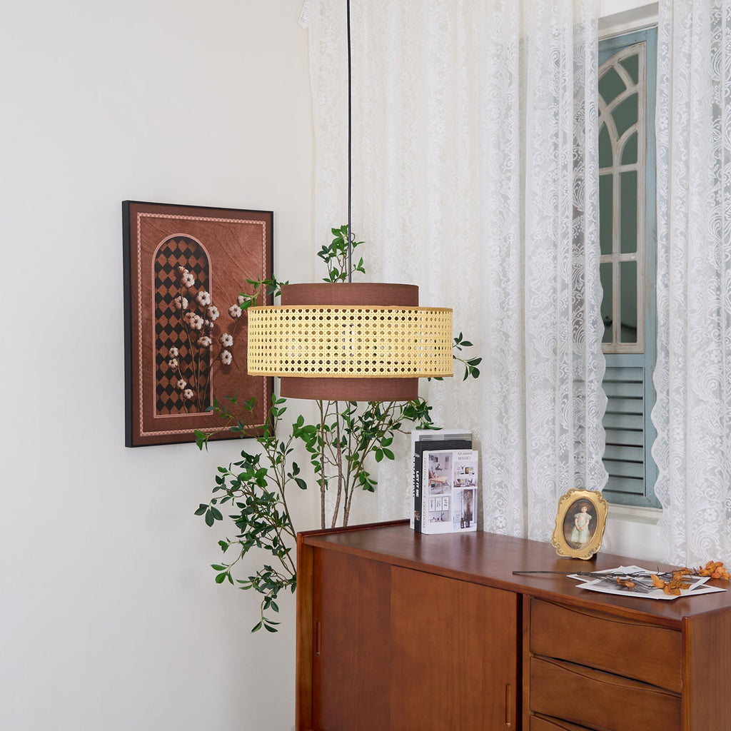 A rattan Clérachic pendant hangs above a wooden sideboard, beside a framed print, lace curtains and a green branch.