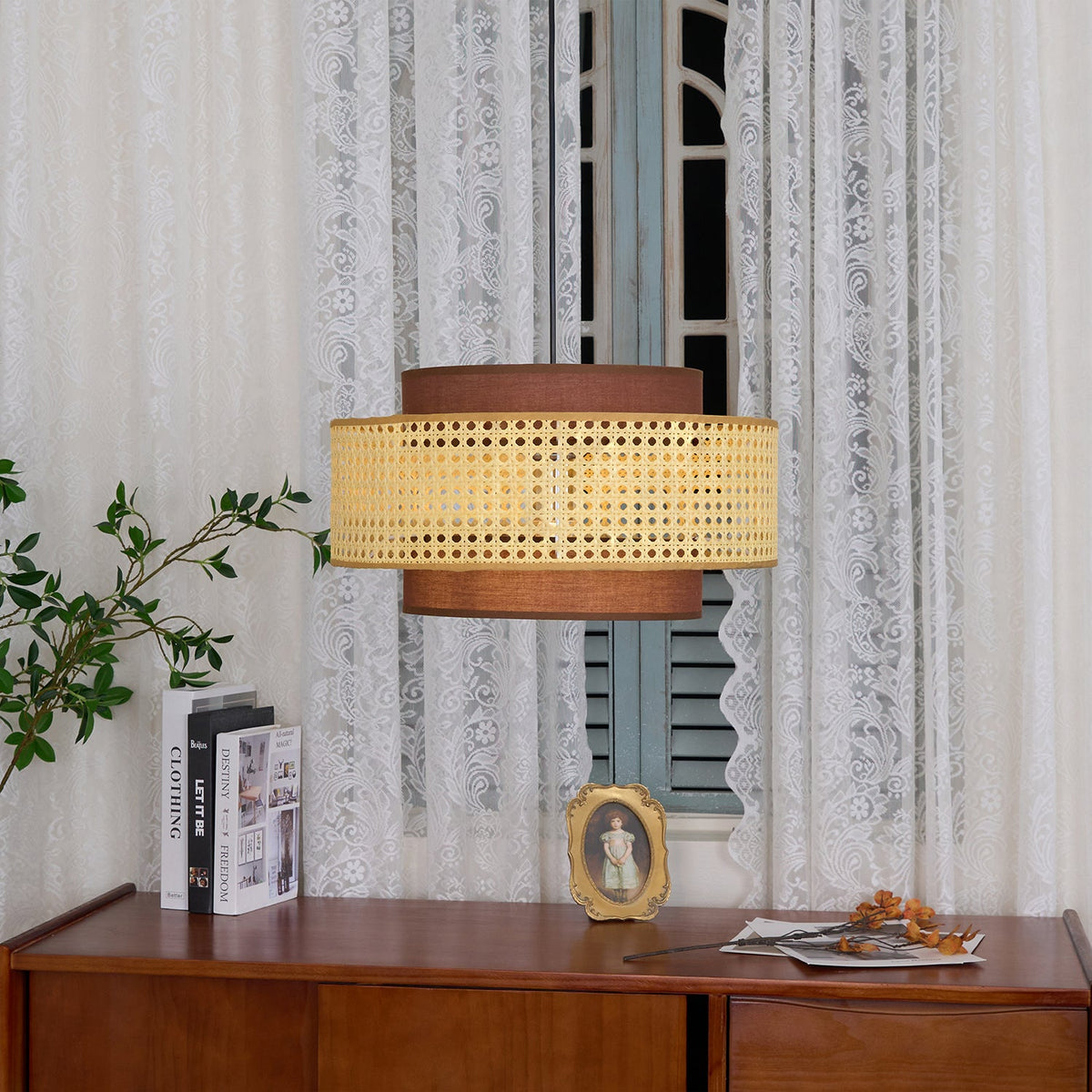 A rattan Clérachic pendant hangs against lace curtains, above two books and a small gold-framed portrait.