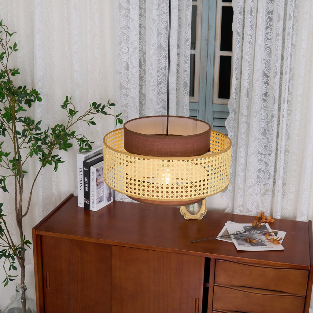 A rattan Clérachic pendant hangs low above a wooden cabinet, with its brown fabric top tier showing and a tall plant beside it.
