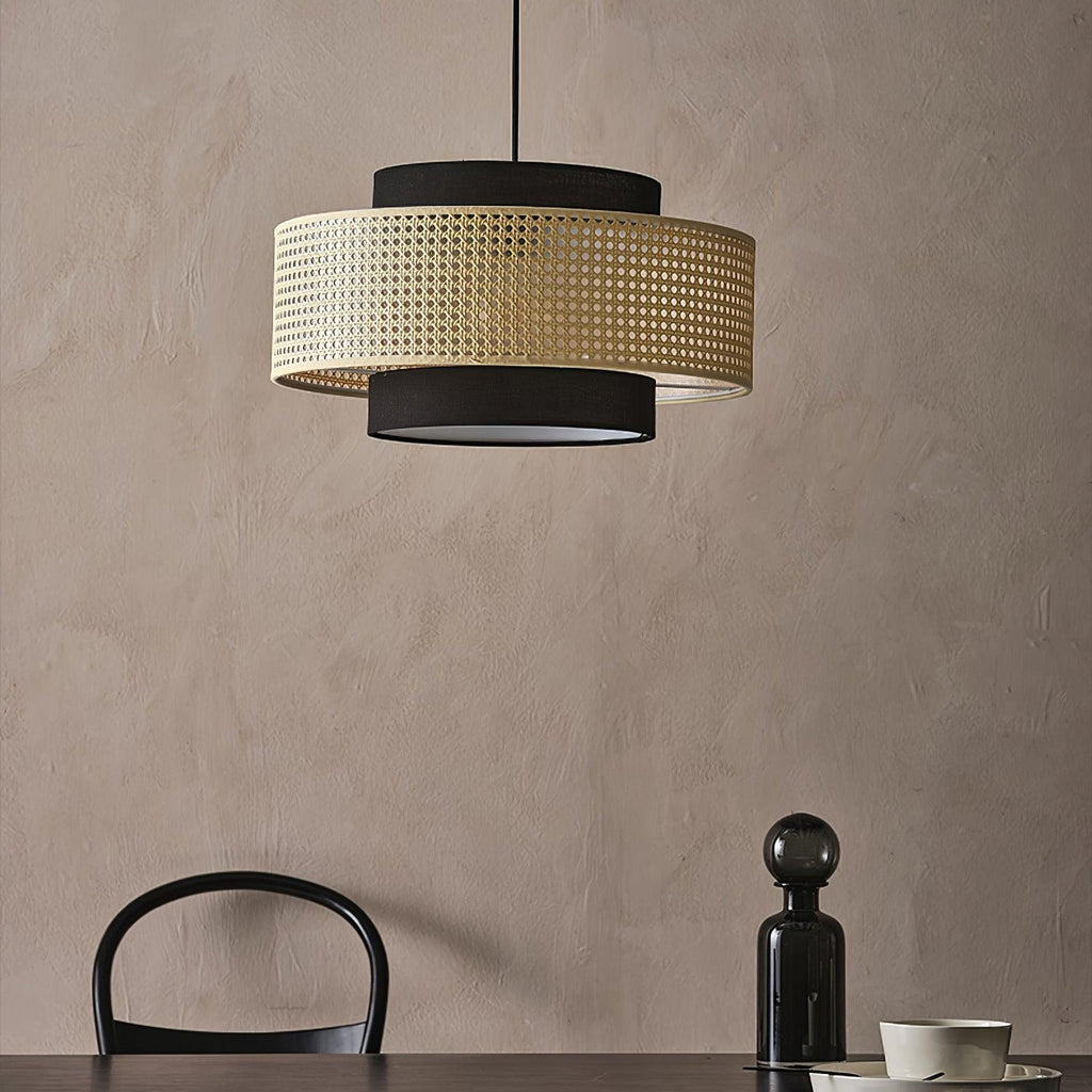 A Clérachic pendant with a rattan drum and black fabric tiers hangs against a clay-pink wall, above a black glass bottle and a chair back.