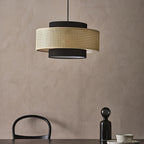 A Clérachic pendant with a rattan drum and black fabric tiers hangs against a clay-pink wall, above a black glass bottle and a chair back.