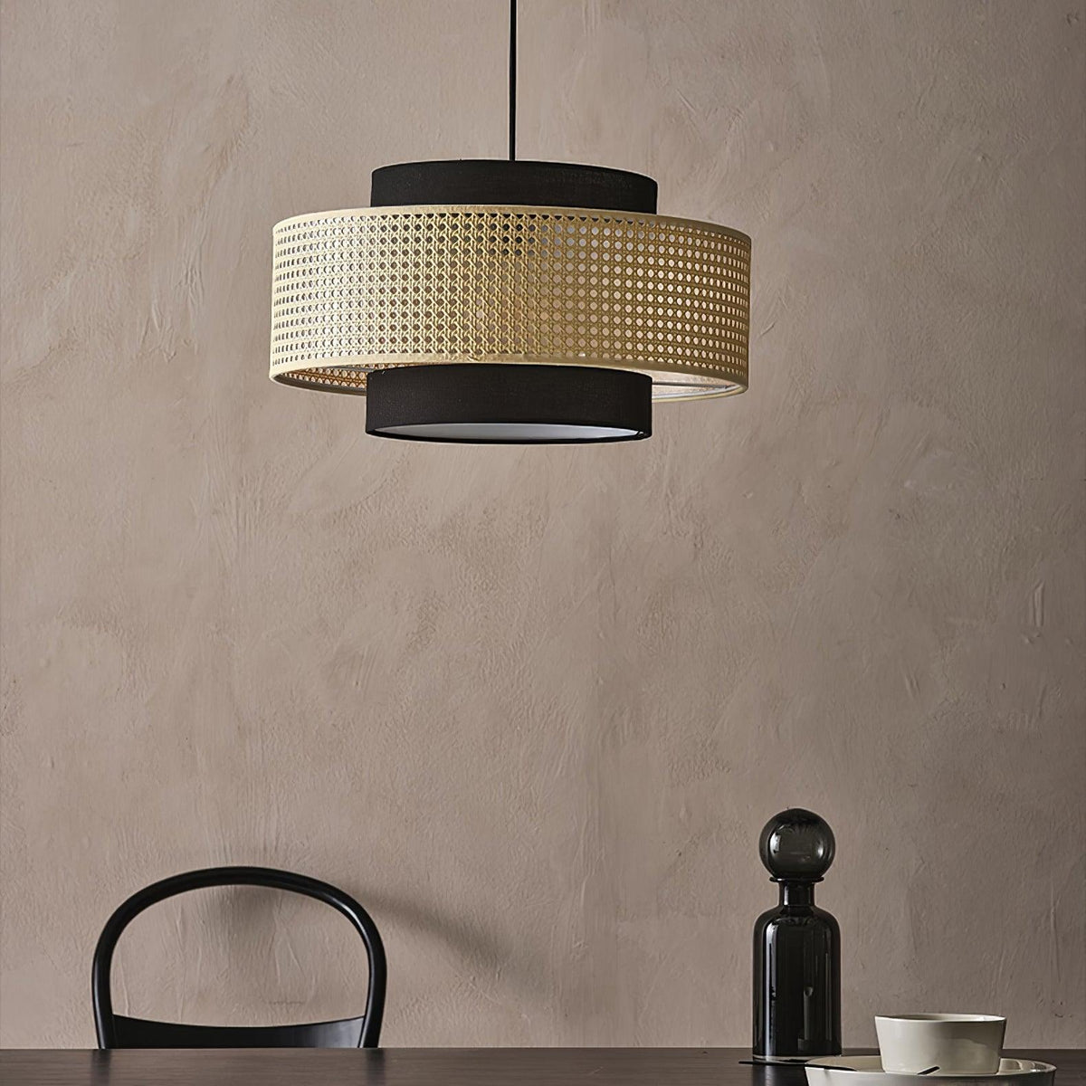A Clérachic pendant with a rattan drum and black fabric tiers hangs against a clay-pink wall, above a black glass bottle and a chair back.