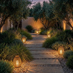 Four Clérachic slatted lanterns glowing at dusk along a flagstone garden path lined with olive trees and grasses.