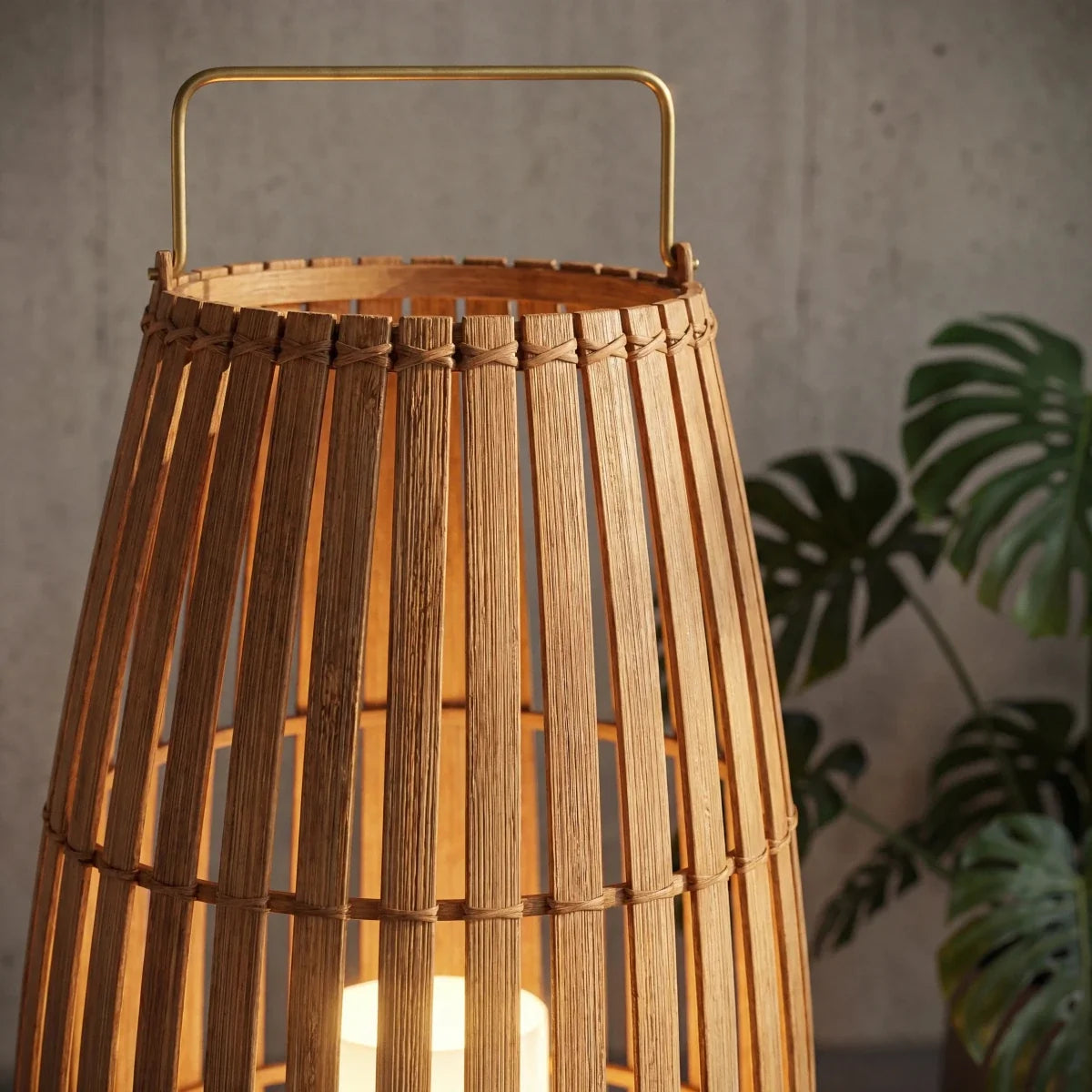 Close view of Clérachic slatted lantern with brass-toned handle and bound rim, set against a concrete wall and monstera leaves.