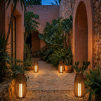 Four tall slatted lanterns glowing at dusk along a stone courtyard path among agave, cactus and ferns, Clérachic outdoors.