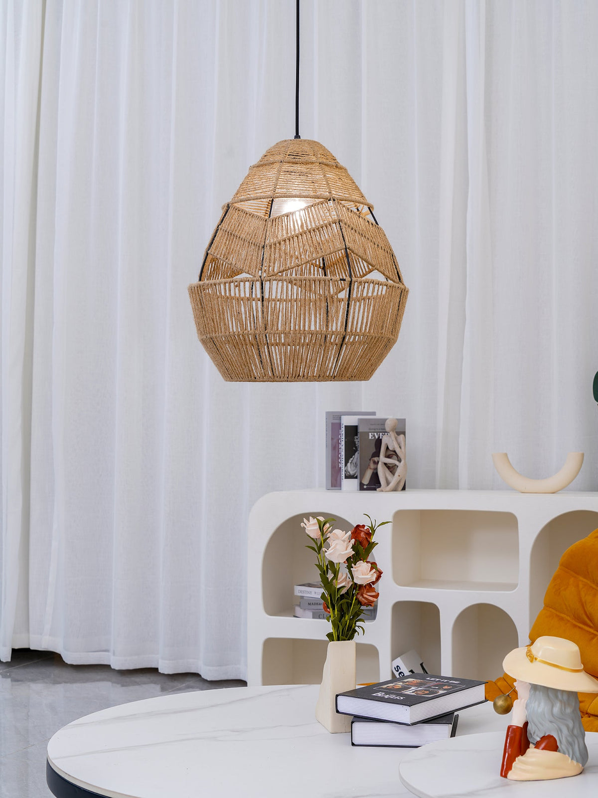 A Clérachic rattan teardrop pendant hangs against white curtains above a marble table with flowers and books.