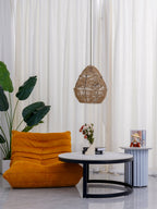 A Clérachic woven pendant hangs in a bright corner with a plant, an orange lounge chair and a low round table.