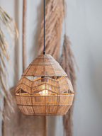 Natural Clérachic rattan pendant lit from inside, hanging in front of dried pampas grass on a grey wall.