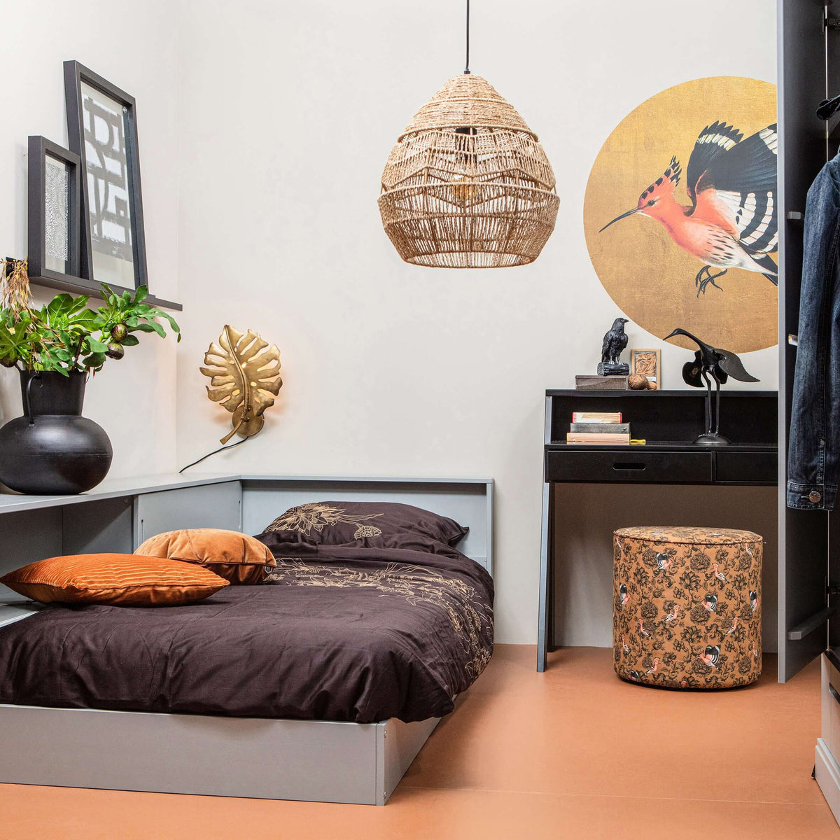 A natural Clérachic rattan pendant hangs in a bedroom with a dark bed cover, a black desk and a bird artwork.