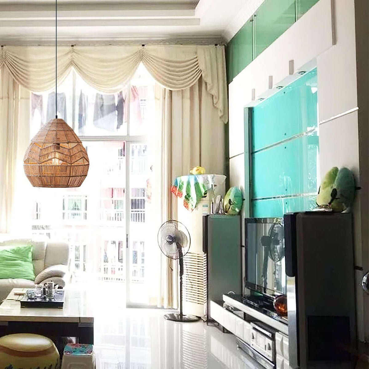 Warm-lit Clérachic rattan pendant in a living room with cream curtains, a glass cabinet and a television.