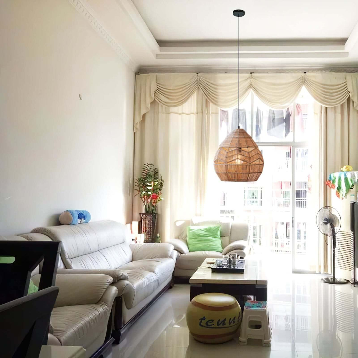 A Clérachic rattan pendant hangs at a living-room window with pleated curtains, a cream sofa and a low table.