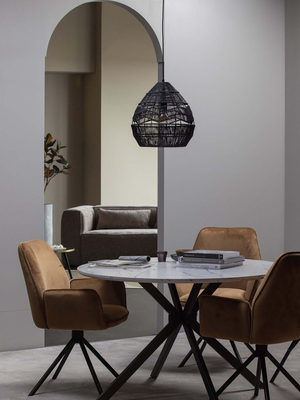 A black Clérachic woven pendant hangs under an arch over a round table with tan velvet chairs.