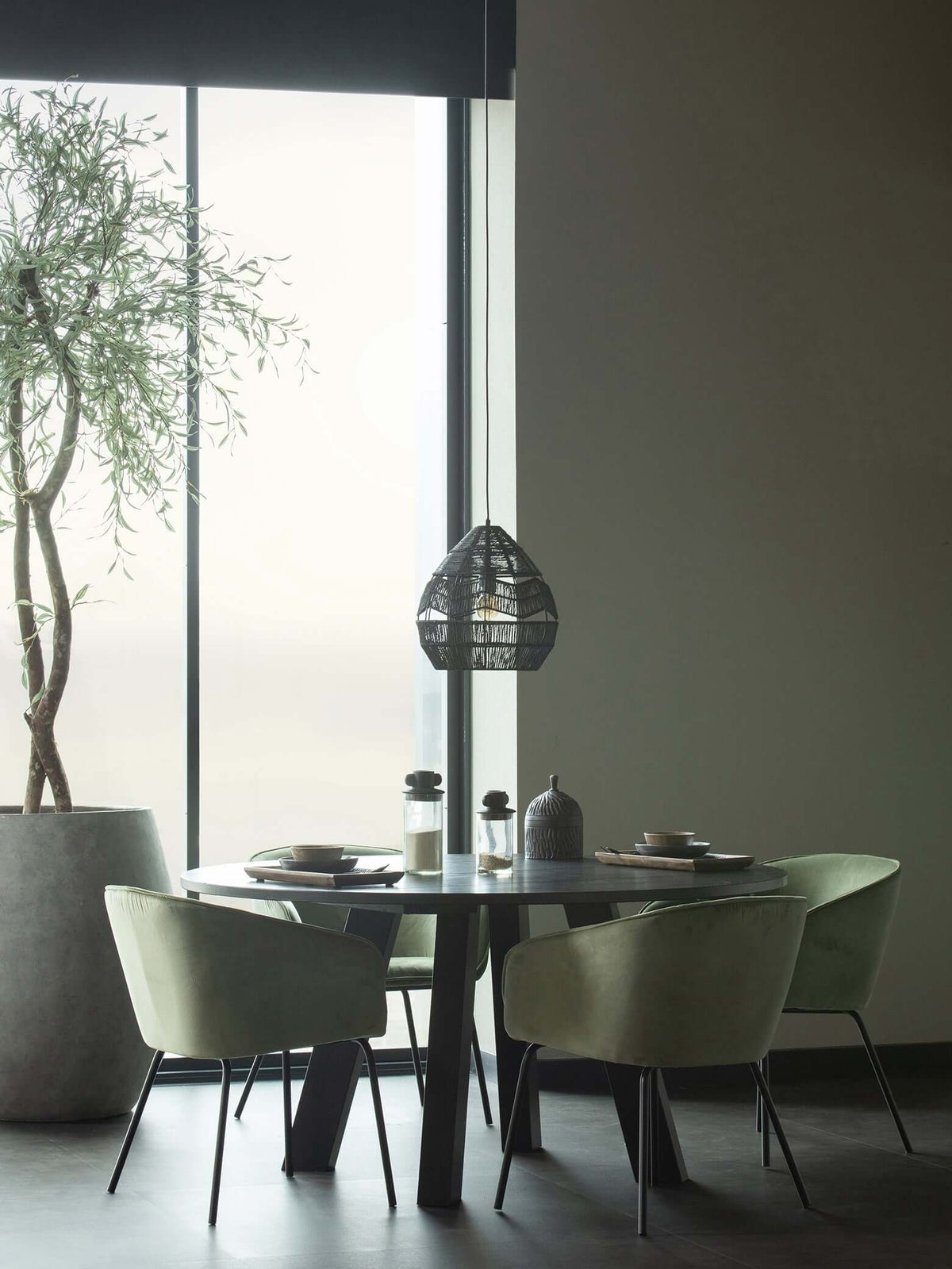 A black Clérachic woven pendant hangs far off over a round dining table with green chairs, by a tall window.