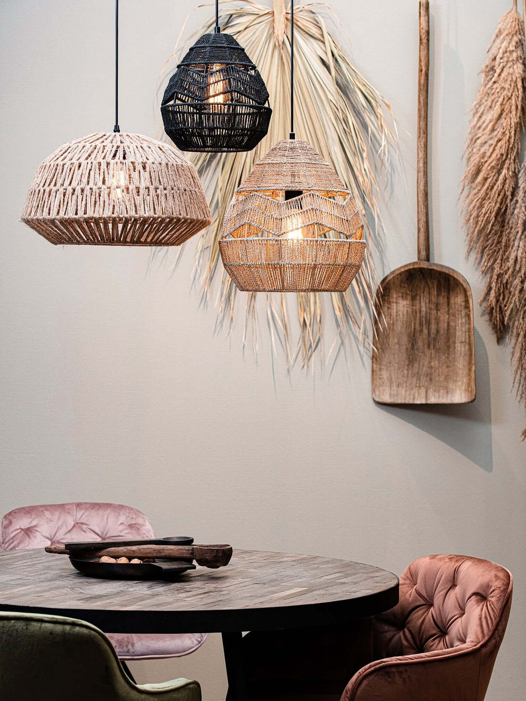 Black and natural Clérachic woven pendants hang beside a third rattan shade, above a dark round table with pink chairs.
