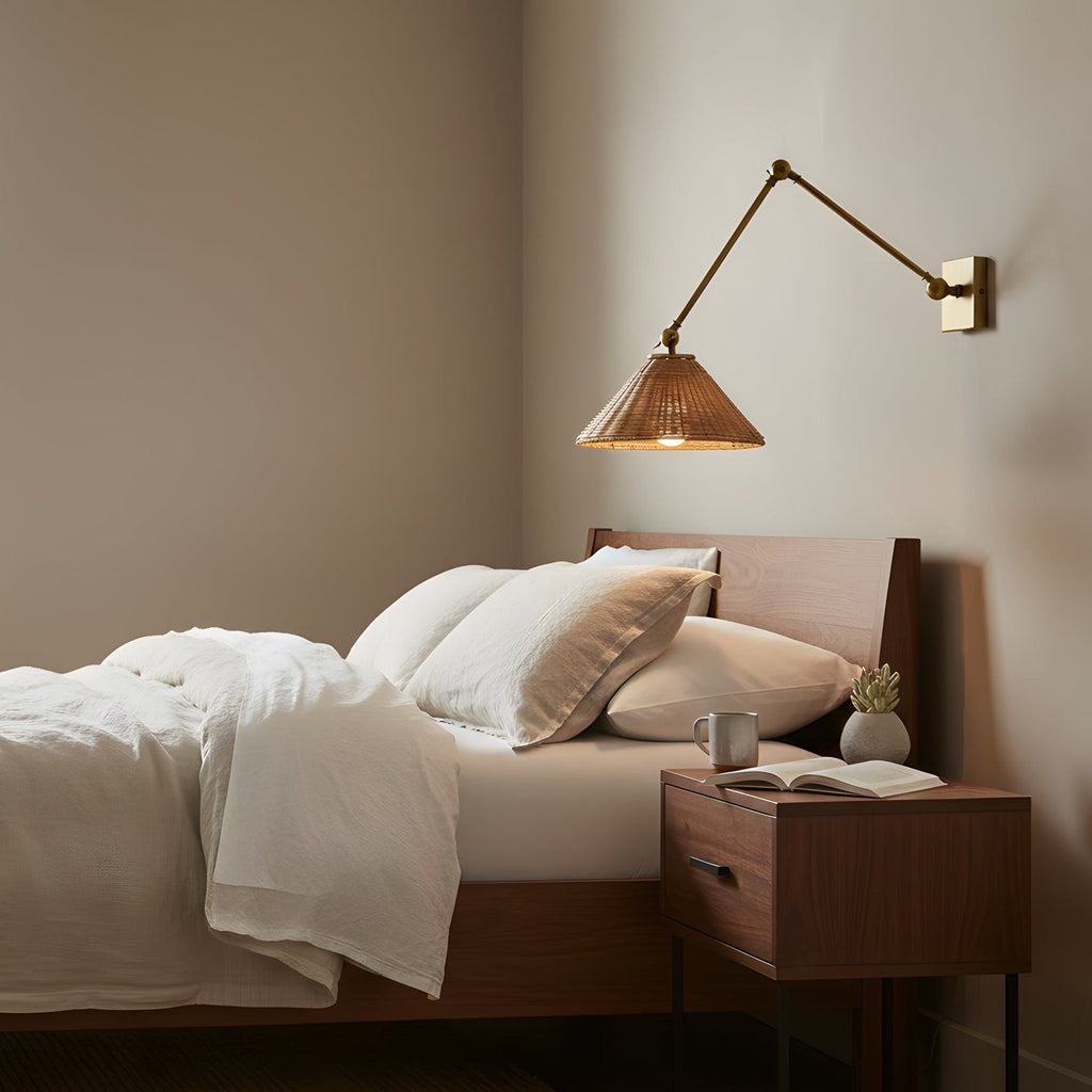 Over a walnut bedside table and a bed with white linen, a rattan-shaded wall light on a gold arm reaches out from a warm wall, from Clérachic.