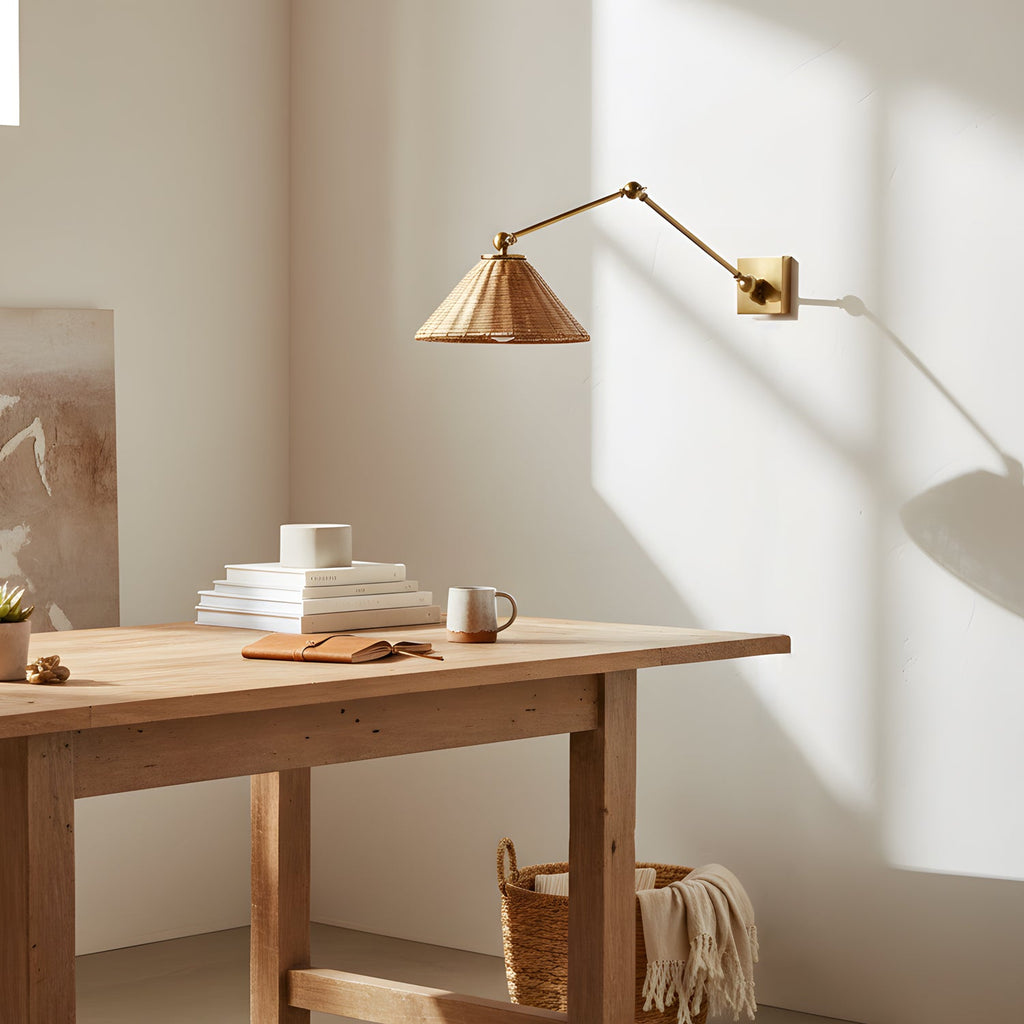 A Clérachic rattan cone wall light on its gold arm catches the sun above a pale wooden desk with stacked books and a mug.