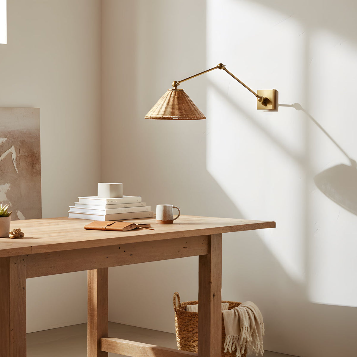 A Clérachic rattan cone wall light on its gold arm catches the sun above a pale wooden desk with stacked books and a mug.