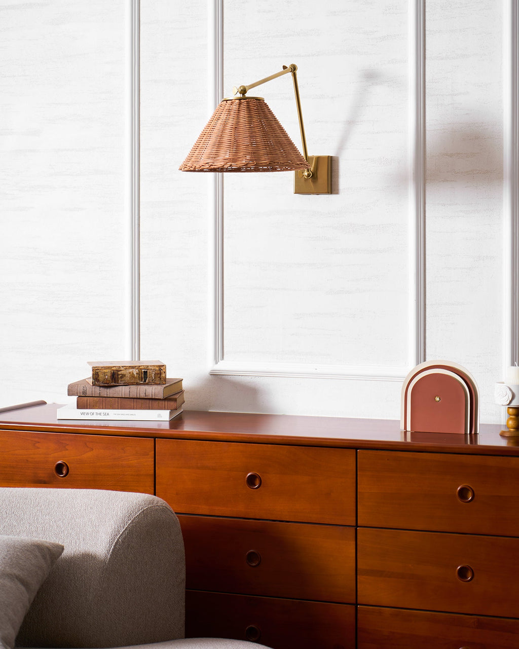 The arm of this Clérachic rattan wall light sits angled to the right, above a wooden chest with books and a small lamp.