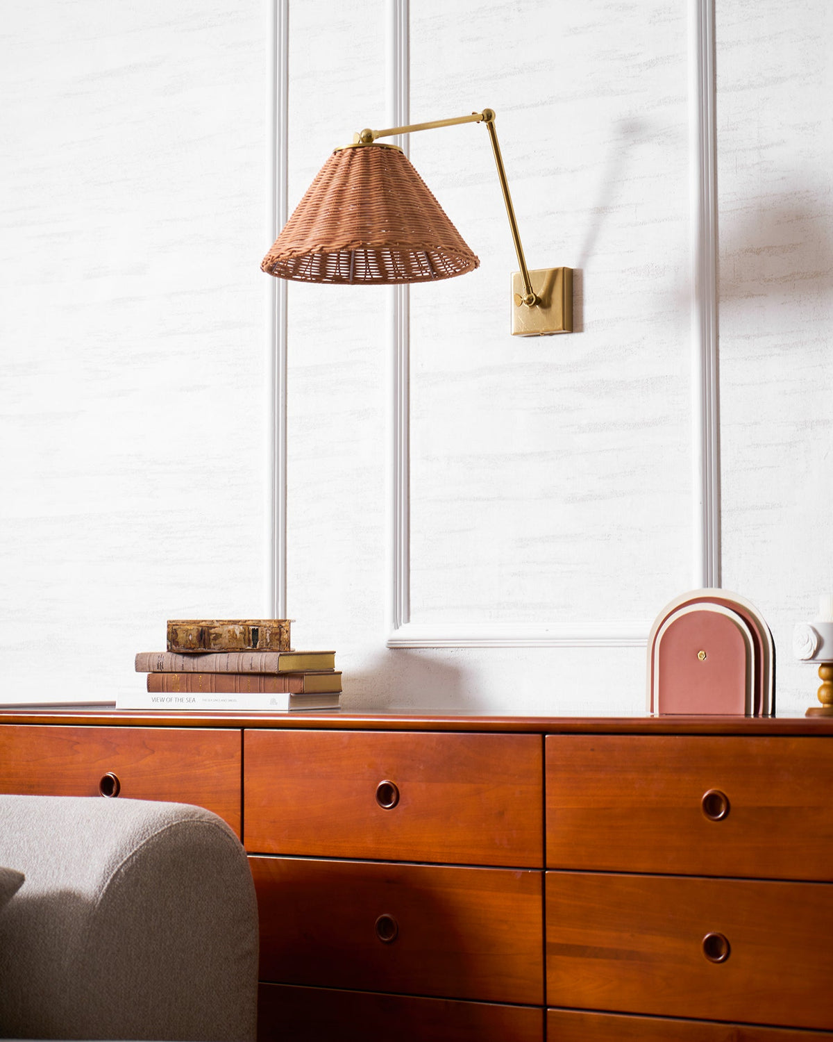 A Clérachic rattan wall light with a gold arm angled to the right of its plate, above a chest of drawers, books and a terracotta arch.