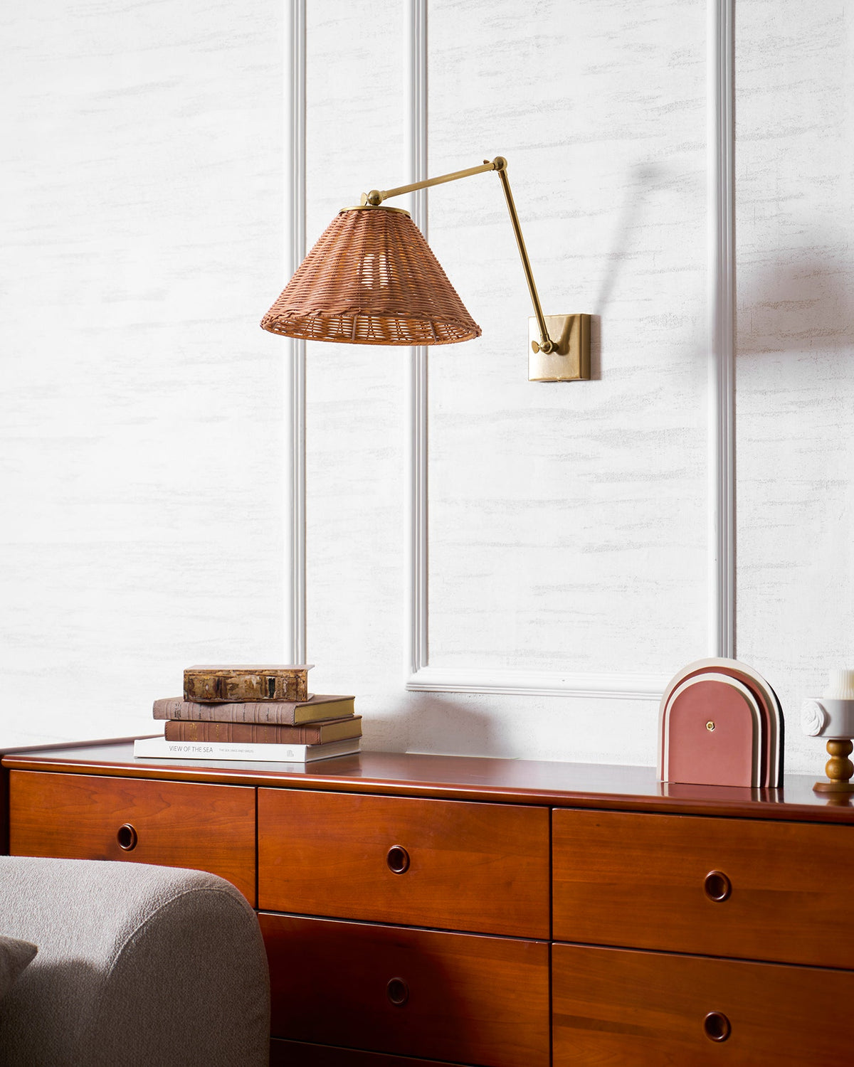 A woven rattan shade on a bent gold arm, over a wooden chest with stacked books and a terracotta arch ornament, from Clérachic.