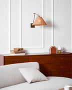 Seen from a distance above a walnut chest and a grey sofa, this Clérachic rattan wall light's gold arm bends toward the wall.