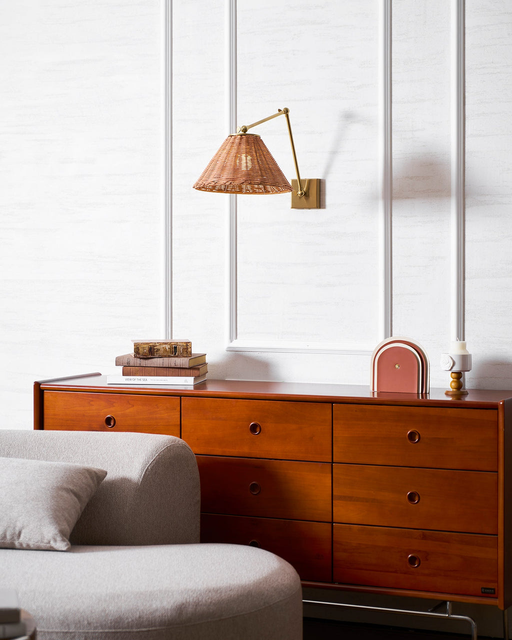 A wider room view of this Clérachic rattan wall light on a white panelled wall, above a drawers unit and a curved grey sofa.