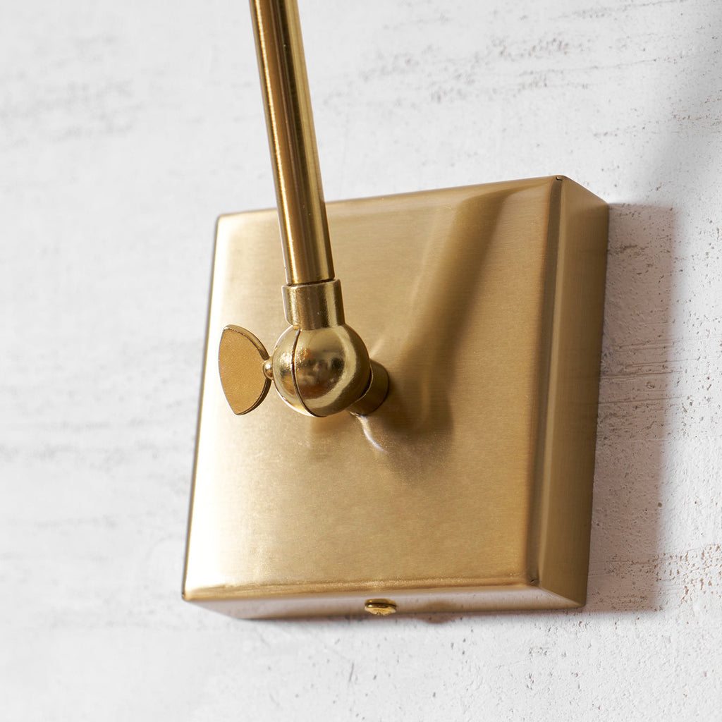 Close view of the square gold wall plate and the pivot knuckle of this Clérachic wall light, on a textured white wall.