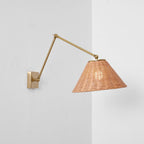 A rattan-shaded Clérachic wall light on a gold arm, hung close to a wall corner, with its bulb glowing faintly inside the weave.