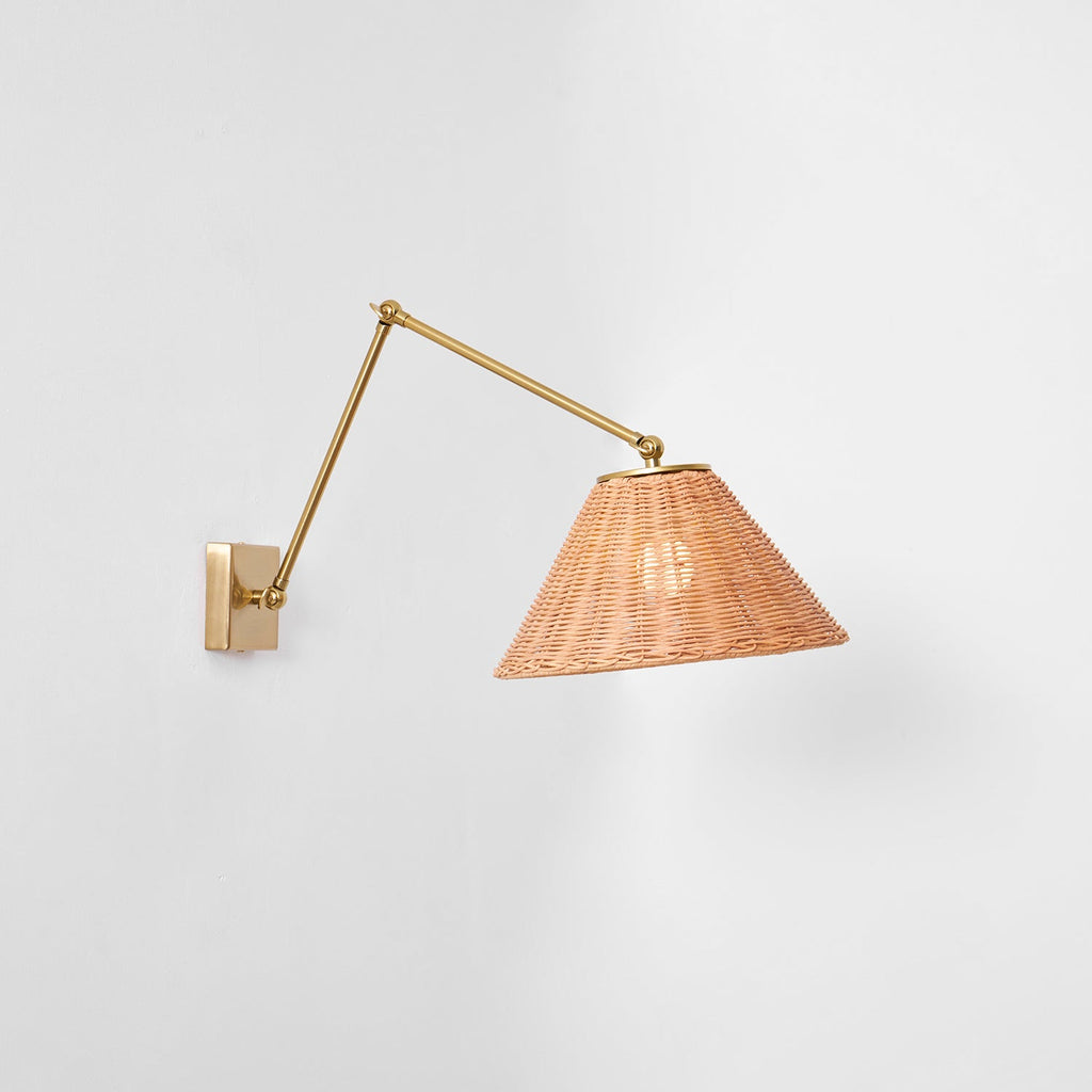 Straight-on view of this Clérachic wall light, with a long gold arm reaching out to a woven rattan cone shade on a pale wall.