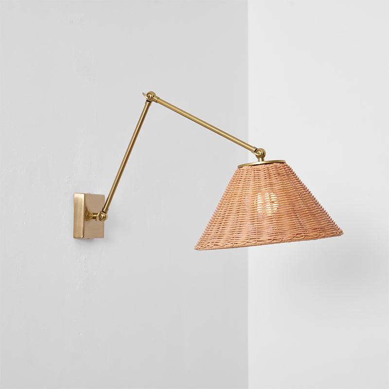 A rattan-shaded wall light on a gold arm, set against a wall corner with a soft shadow, from Clérachic.