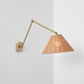 A rattan-shaded wall light on a gold arm, set against a wall corner with a soft shadow, from Clérachic.