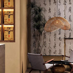 Clérachic rattan pendant in a tea room with a calligraphy wall, tall bamboo plants and woven lounge chairs beside it.