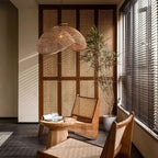 Lit Clérachic rattan wave pendant hanging in a corner room with a rattan cabinet, a low round table and a tall potted tree by the blinds.