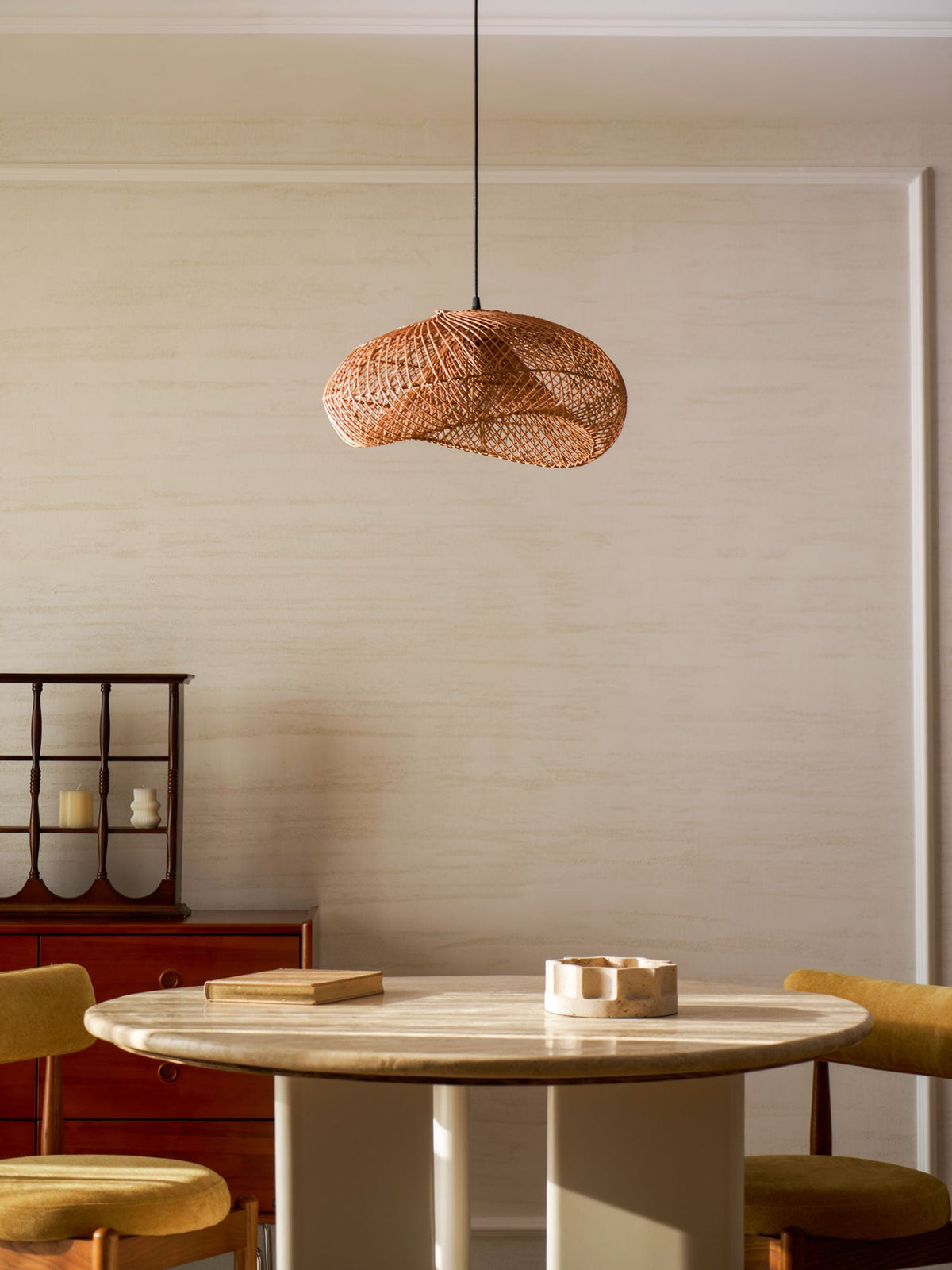 Smaller Clérachic rattan wave pendant over a round table with mustard chairs and a red cabinet, in a quiet cream dining corner.